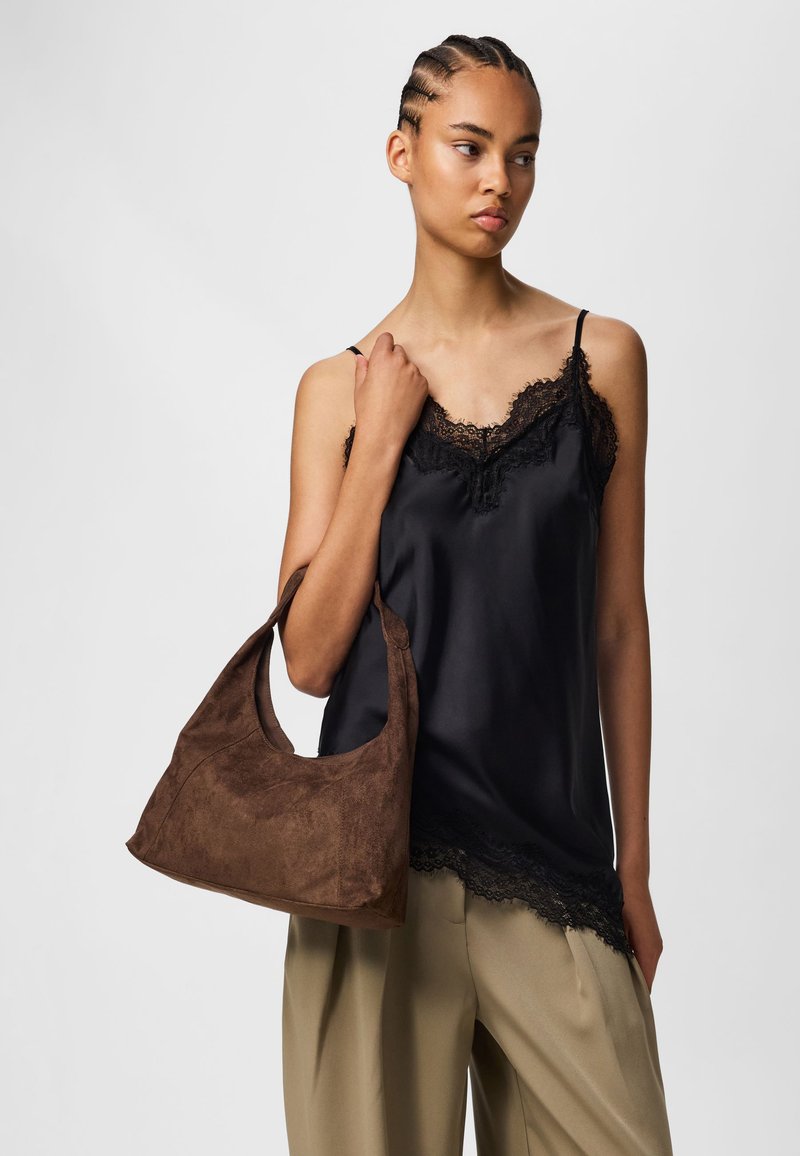 Femme aux cheveux tressés portant un caraco en satin noir bordé de dentelle, un pantalon beige à jambes larges, et portant un sac à bandoulière en daim marron sur le bras droit.
