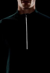 Vyras, užsivilkęs tamsiai mėlyną džemperį su užtrauktuku, kuris iš dalies užtrauktas, prieš juodą foną, matomas tik apatinis veidas ir kaklas.