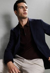 Blazer blu navy sopra un polo bordeaux scuro, pantaloni color crema. Tessuto liscio con vestibilità su misura e cuciture dettagliate. Collo leggermente rialzato sul polo.
