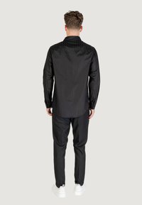 Camicia nera con bottoni, caratterizzata da un motivo testurizzato sulle spalle, abbinata a pantaloni neri e sneakers bianche. Mostrata di spalle.