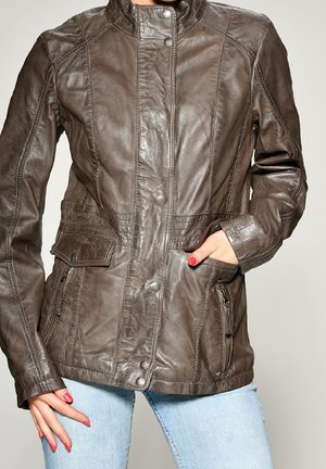 Chaqueta de cuero marrón con botones de presión, bolsillos con cremallera y solapa, usada por una persona con la mano en el bolsillo y uñas pintadas de rojo.