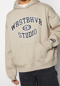 Beige hoodie met een voorvak, met in zwarte, dikke letters "WRSTBHVR" en "STUDIO". Zachte stof, ontspannen pasvorm en geribbelde mouwen.