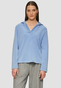 Lichtblauwe hoodie met een V-hals en lange mouwen, met een zachte stoftextuur en een losse pasvorm. Gecombineerd met grijze broeken.