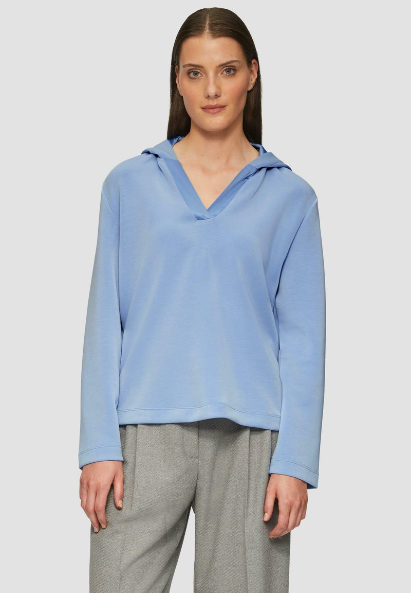 Lichtblauwe hoodie met een V-hals en lange mouwen, met een zachte stoftextuur en een losse pasvorm. Gecombineerd met grijze broeken.