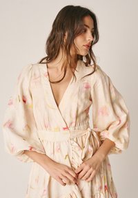 Vrouw met los bruin haar, gekleed in een crèmekleurige bloemenwikkeljurk met pofmouwen, kijkt opzij, handen in elkaar gevouwen ter hoogte van de taille.