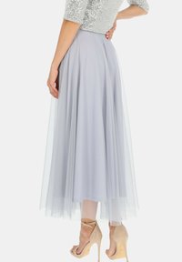 Jasnoszara, sięgająca kolan spódnica z tiulu o zwiewnym fasonie. Modelka ma na sobie beżowe szpilki. Widoczny błyszczący srebrny top.
