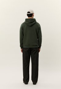 Muž v tmavozelenej mikine s kapucňou, čiernych džínsach, čiernych topánkach a béžovej šiltovke stojí chrbtom k divákovi na jednoduchom pozadí.