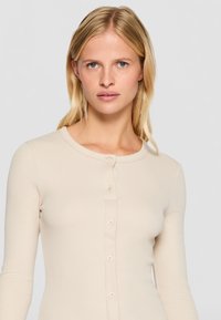 Beige långärmad topp i ribbat material med rund halsringning, prydd med knappar framtill och en figursydd silhuett.