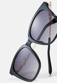 Occhiali da sole neri oversize con lenti grigie sfumate, forma angolare distintiva, sottili dettagli in metallo color oro rosa sulle stanghette e una finitura lucida.