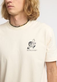 Camiseta de algodón color beige claro con un gráfico negro de una tortuga y el texto "REVOLUTION™" en una fuente casual en el área del pecho izquierdo.