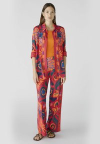 Rode en oranje bloemenprint outfit met een knoopsluiting overhemd en bijpassende wijde broek, gecombineerd met een oranje top en zwarte sandalen.
