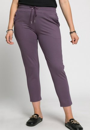 Woman wearing purple drawstring pants with ruched pockets and black slip-on loafers with gold chain detail, standing against neutral background.