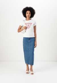 T-shirt bianco con stampa grafica rossa, abbinato a una gonna di jeans blu. Sandali bianchi e una piccola borsa beige completano il look.