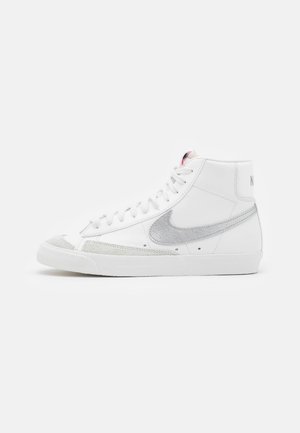 Witte hoge sneaker met een zilveren Nike-swoosh, witte veters en lichtgrijze suède accenten op de neus en zijkanten.
