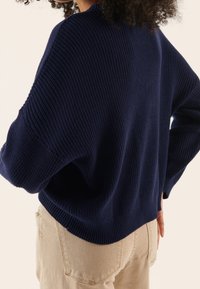 Granatowy sweter z dzianiny o prążkowanej fakturze, z szerokimi rękawami i luźnym krojem. W połączeniu z jasnobeżowymi spodniami, prezentującymi neutralną paletę kolorów.