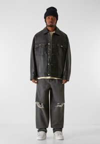 Schwarze Lederjacke in Übergröße mit zwei Fronttaschen, kombiniert mit grauen, zerrissenen Jeans und weißen Sneakers. Getragen mit einer schwarzen Strickmütze.