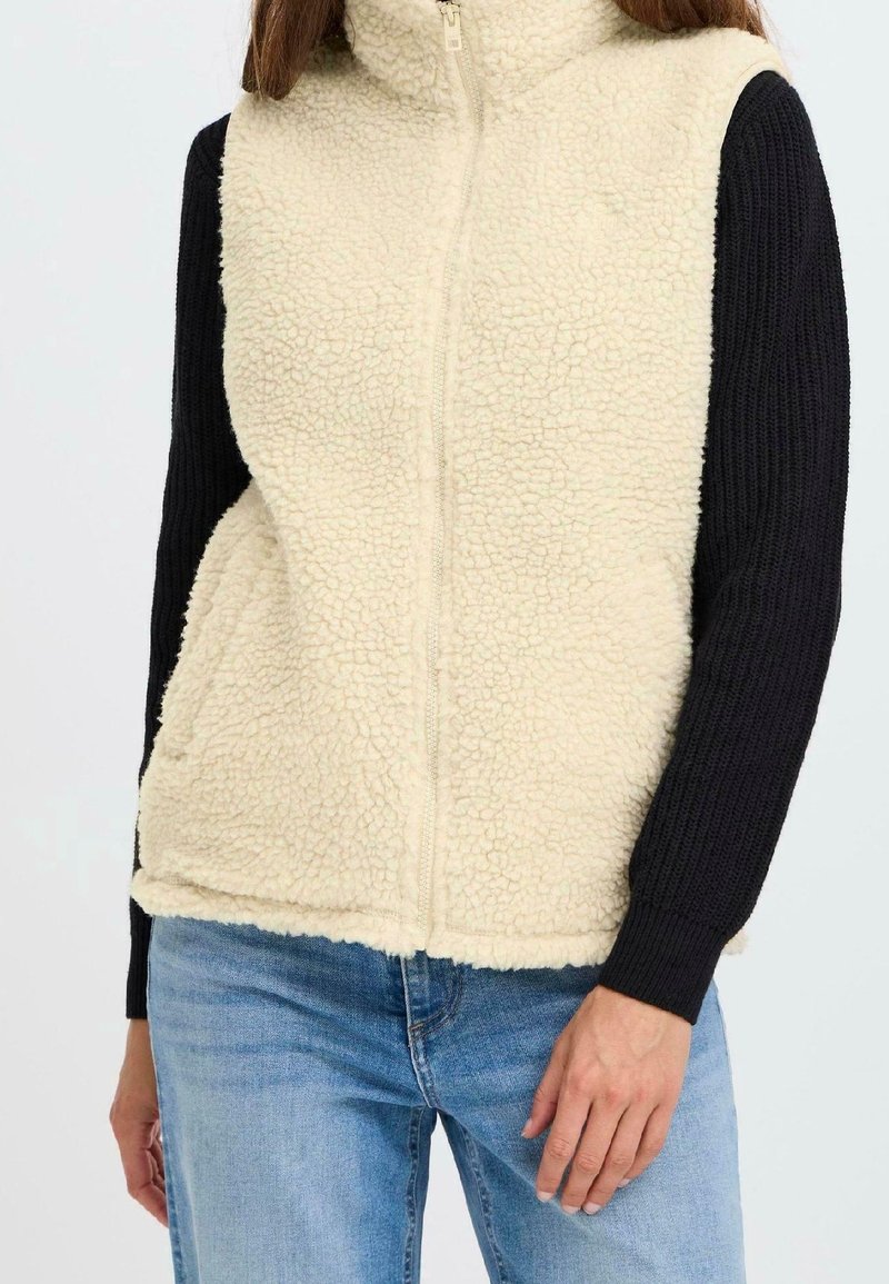 Femme portant un gilet sherpa polaire beige à fermeture éclair sur un pull noir à manches longues et un jean bleu clair, debout avec une main vers le bas.
