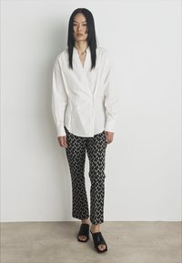 White long-sleeve shirt, wrap design; black patterned trousers with white shapes; open-toe black mules; minimalist aesthetic, neutral background.
