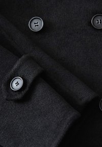 Primo piano di un cappotto di lana nera, con tessuto testurizzato, due bottoni tondeggianti e dettagli del colletto piegato.