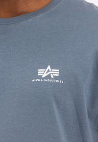 Pelēka T-krekls, kas izgatavots no kokvilnas, ar mazu balto logo, kas sastāv no trim horizontālām līnijām un tekstu "ALPHA INDUSTRIES" krūtīs.