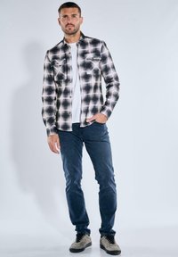 Schwarz-weiß kariertes Flanellhemd mit zwei Brusttaschen, getragen über einem weißen T-Shirt, kombiniert mit blauen Jeans und grauen Sneakers.