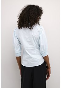 Light blue blouse with puff sleeves, fitted waist, and soft texture. Back features a subtle seam detailing, paired with black trousers.
