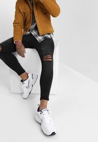 Brązowa kurtka ze sztruksu na czarno-białej koszuli w kratę, do tego czarne, rozdarci skinny jeansy oraz białe buty sportowe z niebieskimi akcentami.