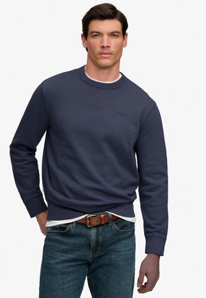 Man wearing navy blue sweatshirt over white shirt, blue jeans, and brown leather belt, standing with one hand in pocket against plain background.