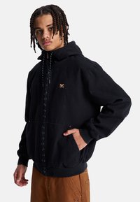 Giacca nera con cappuccio, zip frontale, grandi tasche e toppa con logo. Presenta una vestibilità comoda e cordini regolabili sul cappuccio.