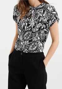 Žena oblečená v černo-bílé košili s květinovým potiskem a krátkým rukávem, zasunuté do černých kalhot, se zdrženlivě dívá s rukama v kapsách.