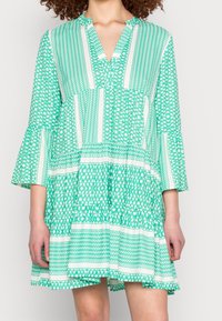 Vestido verde con estampado, escote en V, mangas acampanadas y falda en capas. Presenta varios diseños geométricos y franjas blancas.