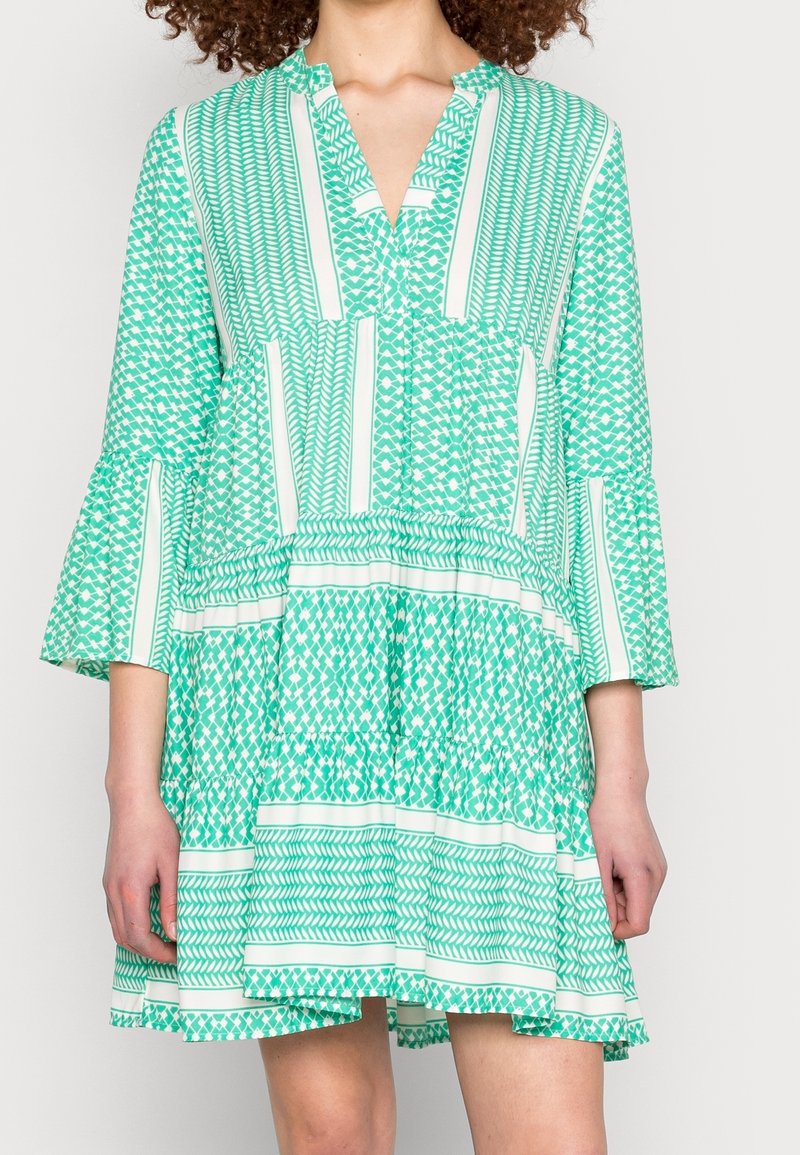 Vestido verde con estampado, escote en V, mangas acampanadas y falda en capas. Presenta varios diseños geométricos y franjas blancas.