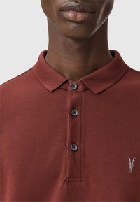 Polo marrone con tessuto testurizzato, dotato di patta con tre bottoni e un piccolo logo ricamato in bianco sul petto.