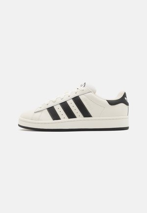Weißer Adidas Sneaker mit schwarzen Streifen und Fersenlasche, niedrig geschnitten, Schnürverschluss und flacher Sohle vor einfarbigem Hintergrund.