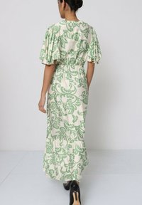 Robe maxi en crème avec un motif floral vert, dotée de manches volantes, d'une taille élastique et d'un ourlet en couches. Tissu lisse et léger.