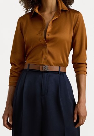 Vrouw draagt een bruine blouse met knopen, ingestopt in een donkerblauwe broek met hoge taille, met een bruine riem met een zilveren RL gesp.