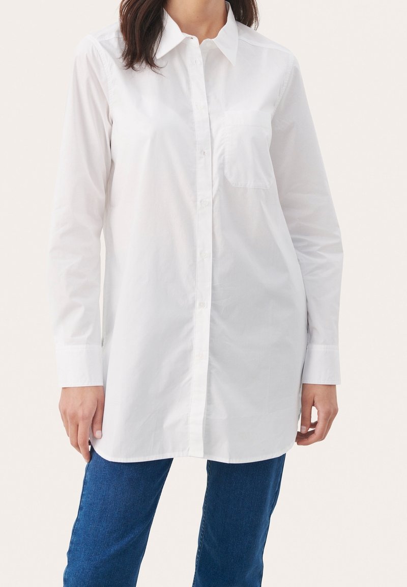 Vrouw in een lange witte blouse met knopen en kraag, voorzien van een borstzak, gecombineerd met een blauwe spijkerbroek tegen een effen lichte achtergrond.