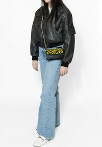 Giacca bomber in pelle nera, jeans a zampa d'elefante blu chiaro, scarpe bianche e una piccola borsa a tracolla a strisce zebra nere e gialle con tracolla a catena.