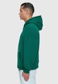 Grøn hættebluse med kængurulomme, ribbede manchetter og en afslappet pasform, vist fra siden mod en neutral baggrund.