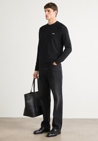 Schwarzes Langarmshirt mit kleinem weißen Logo, kombiniert mit schwarzen Jeans und einer schwarzen Ledertasche. Elegante Schuhe runden das Outfit ab.
