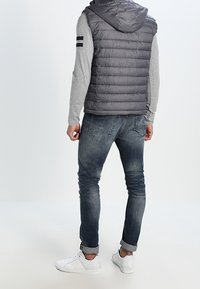Grå pufferväst med huva, bärs över en grå långärmad tröja. Kombinerad med mörka denimjeans och vita sneakers. Enkelt mönster med ränder.