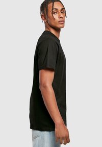 Sort bomulds T-shirt med rund hals og korte ærmer, der viser et tætsiddende design. Modellen poserer sidelæns mod en lys grå baggrund.