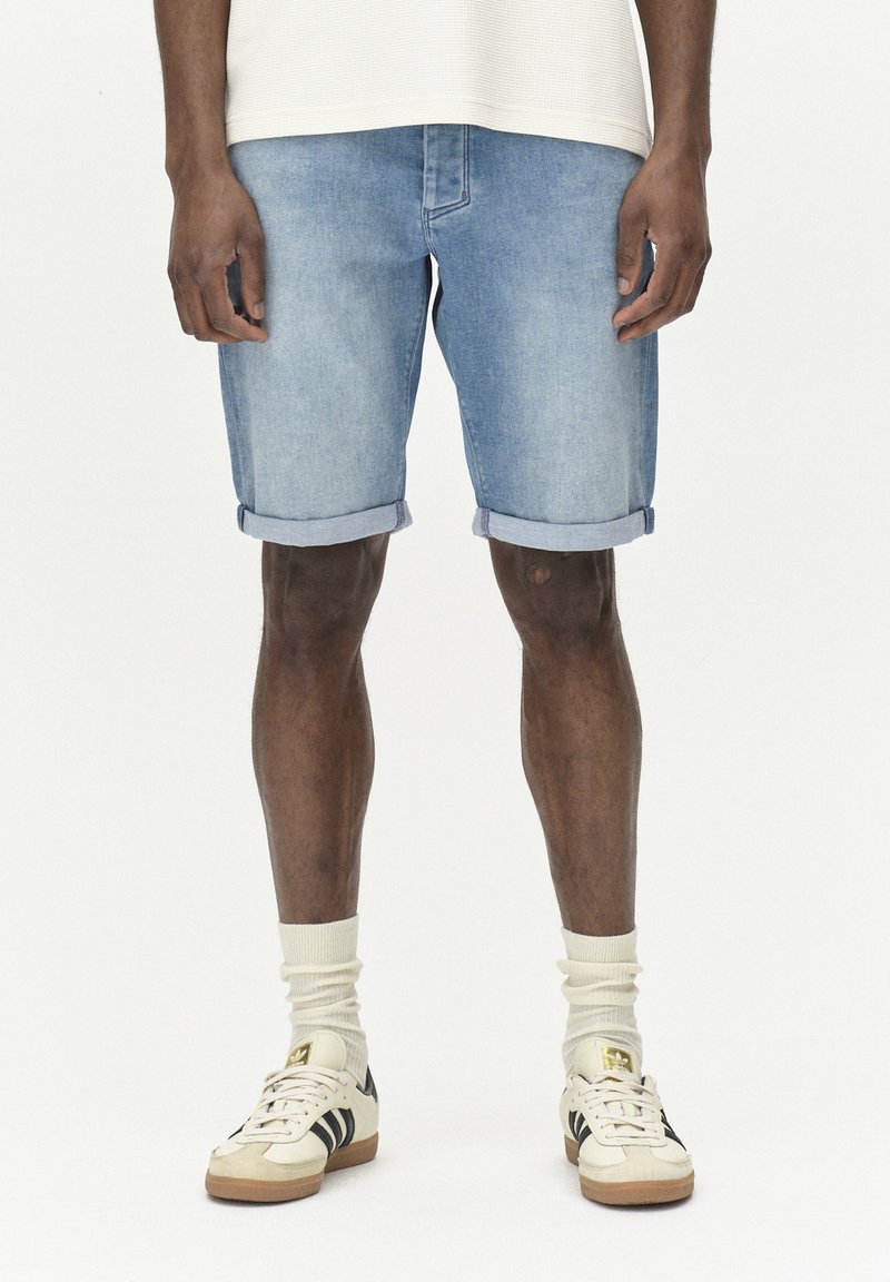 Shorts de mezclilla azul claro con dobladillos enrollados, hechos de algodón, combinados con zapatillas beige y calcetines color crema. Diseño simple y casual.