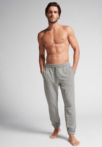 Grijze sweatpants met elastische tailleband en boorden, gemaakt van zachte stof. Model staat blootsvoets, torso bloot, op een neutrale achtergrond.