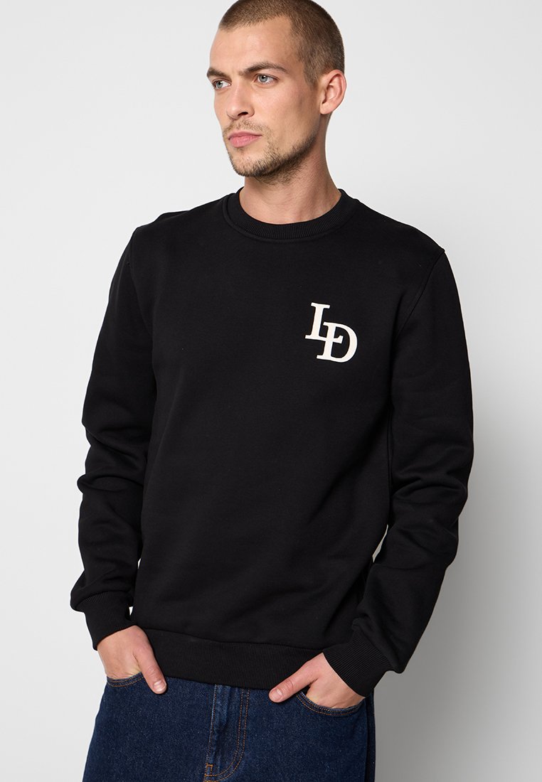 Mann in einem schwarzen Sweatshirt mit weißen "LD"-Buchstaben auf der Brust, blauen Jeans, steht mit den Händen in den Taschen und schaut zur Seite.