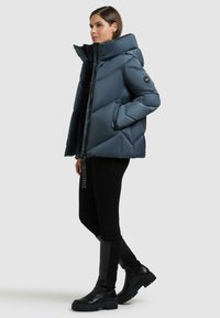 Giacca puffer blu scuro con colletto alto, caratterizzata da un motivo a trapunte, zip frontale e tasche laterali, indossata con leggings neri e stivali.