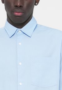 Ανοιχτό μπλε πουκάμισο με κλασικό γιακά, μία τσέπη στο στήθος και λείο βαμβακερό ύφασμα. Τα λευκά κουμπιά δημιουργούν αντίθεση με το ύφασμα.