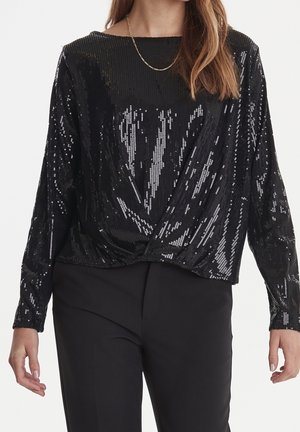 Kobieta ubrana w czarną, długą bluzkę z cekinami z skręconym detalem z przodu oraz czarne spodnie, uzupełniona cienkim złotym łańcuszkiem na szyi.