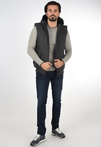 Svart hettevest over en lys grå langermet skjorte, kombinert med mørke jeans og grå sneakers. Har glidelås og lommer.
