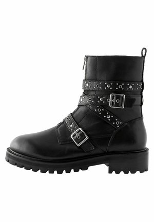 Black leather ankle boot with buckles and studded straps, round toe, and chunky tread sole. Side zipper for easy access.